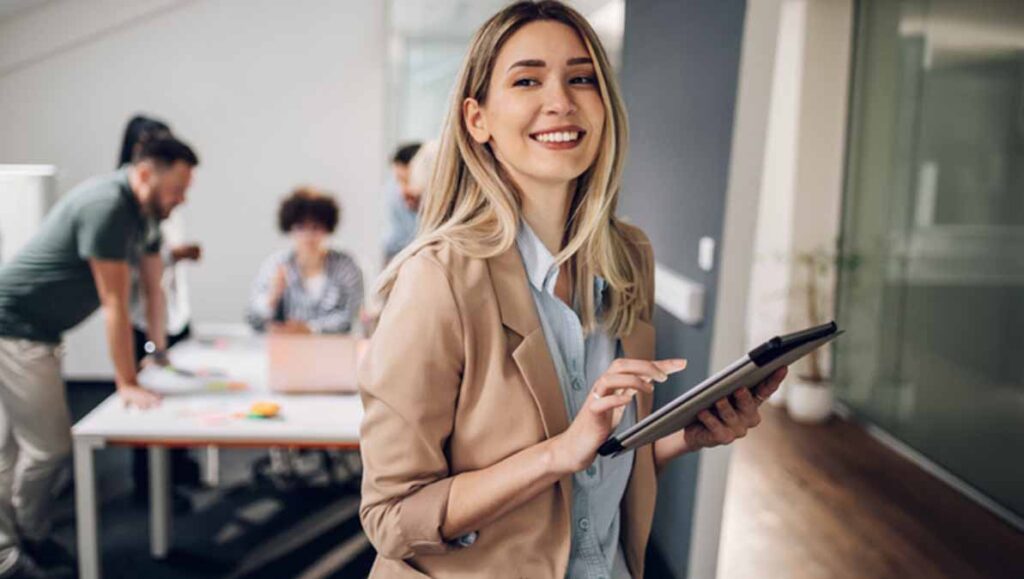 A young business casual Caucasian women holding a tablet while smiling just beyond the camera with a group of people crowded at a desk behind her, working and discussingFrom Tech to Transformation: Why People and Process Matter in Tech Initiatives